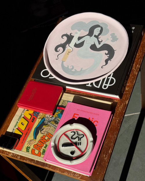 A curation of coffee table books, a vintage comic, a baby pink, ceramic tray and a No Smoking Ashtray on an old wooden surface.