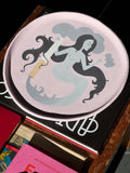 A curation of coffee table books, and a baby pink, hand-painted, ceramic tray on an old wooden surface.