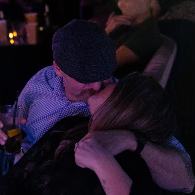 A couple embracing and kissing in a dimly lit, atmospheric venue with purple and blue ambient lighting. The man is wearing a dark flat cap and a light blue patterned shirt, while the woman has long light brown hair and is wearing a black top.