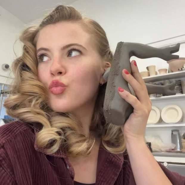 Jessica Booty, a woman with blonde retro-style curls holds a gray ceramic sculpture prop to her ear as if on a telephone, wearing a dark brown corduroy jacket in what appears to be a pottery studio.