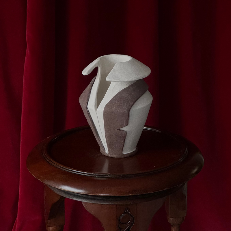 White tuxedo vase ceramic sculpture on a wooden pedestal against a red velvet curtain. A 2025 design by Jessica Booty.