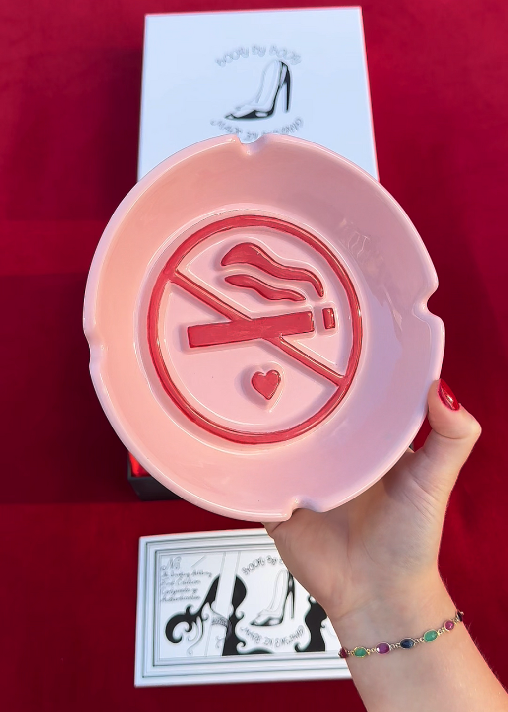 A hand holds a pink ceramic ashtray with a red cigarette ban symbol, positioned in front of its white retail packaging on a red background.