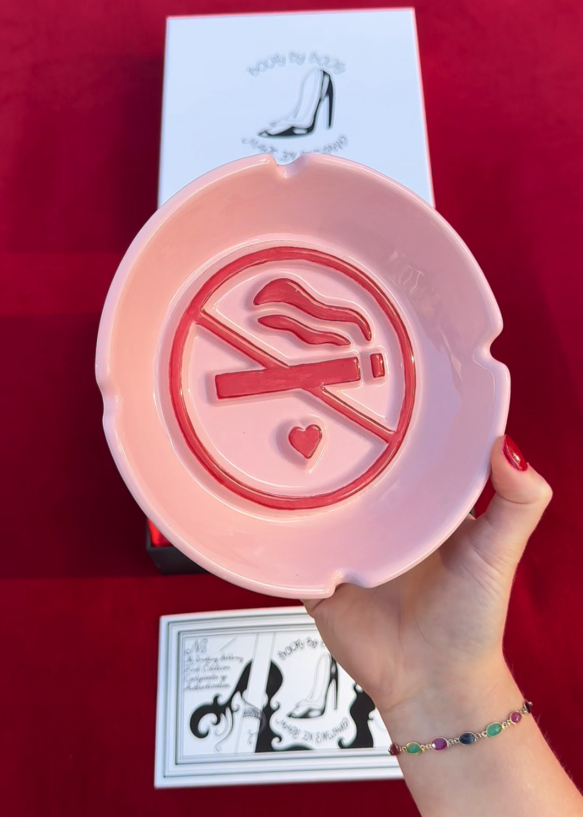 A hand holds a pink ceramic ashtray with a red cigarette ban symbol, positioned in front of its white retail packaging on a red background.