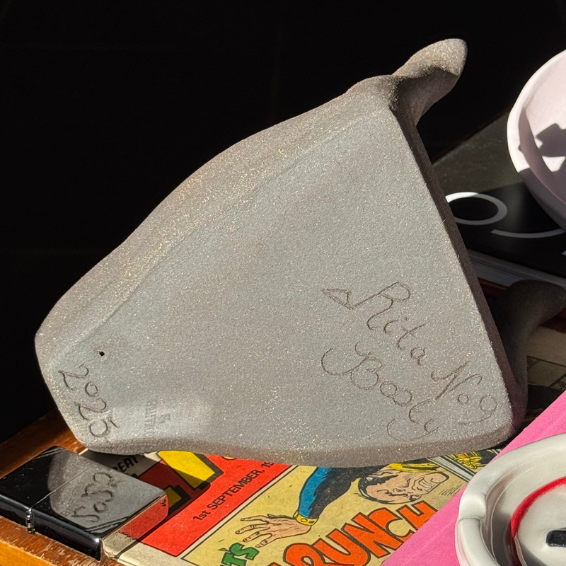The underside of a Risqué Rita ashtray and ceramic sculpture with engraved text on a table with comic book, lighter and other ceramic art.