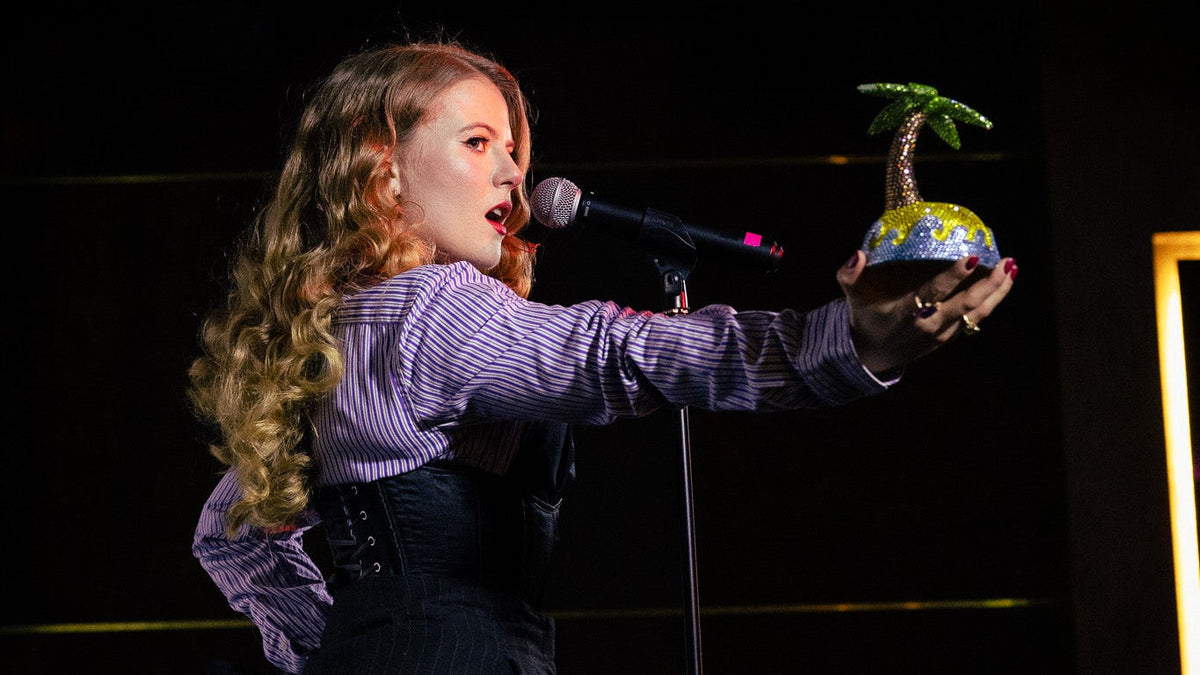 Jessica Booty on stage holding a rhinestoned paradise island ceramic sculpture with palm tree, singing into a microphone.