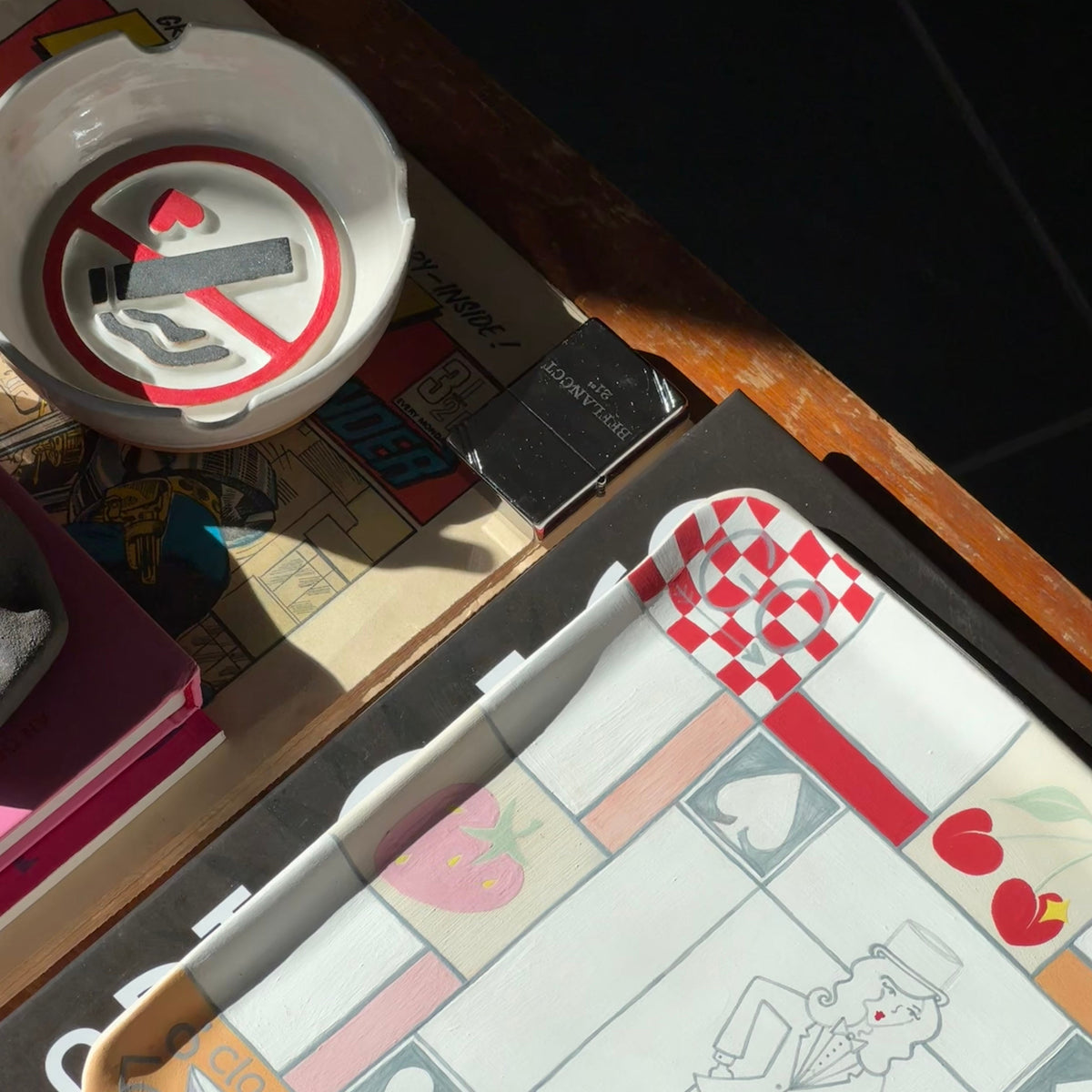 A close-up, angled view of a board game board featuring a whimsical retro design with a line-art character, hearts, cherries, and a red-and-white checkered flag. The board is styled on a wooden table alongside a white ceramic no smoking ashtray and other decorative items.