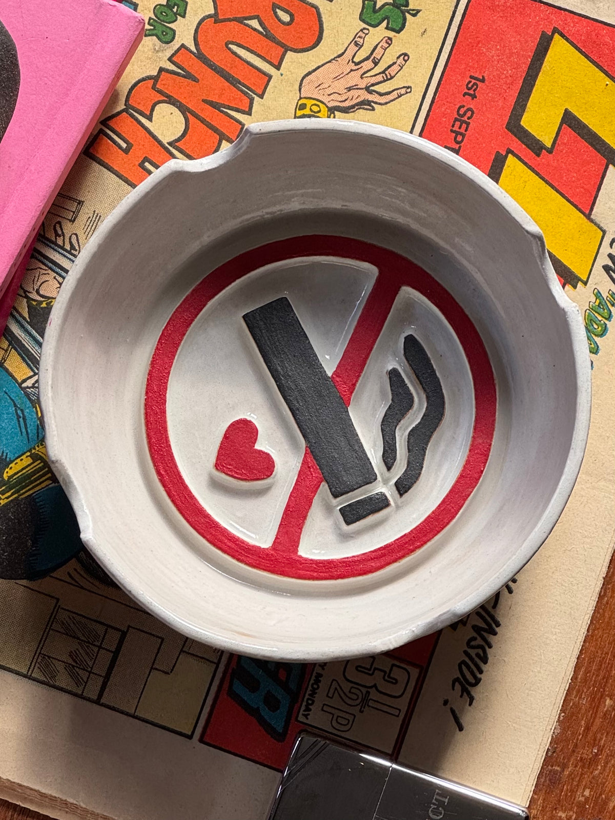 A white ceramic no smoking ashtray featuring a 'no smoking' symbol with a small red heart, shown resting on a colourful vintage comic book on a coffee table.